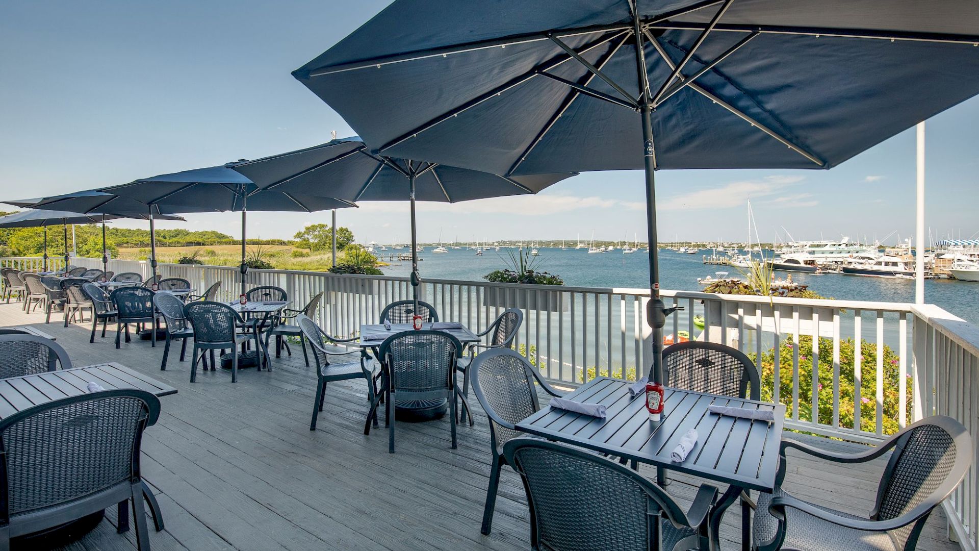 An outdoor dining area with tables, chairs, and blue umbrellas overlooks a scenic waterfront with boats and greenery in the background.