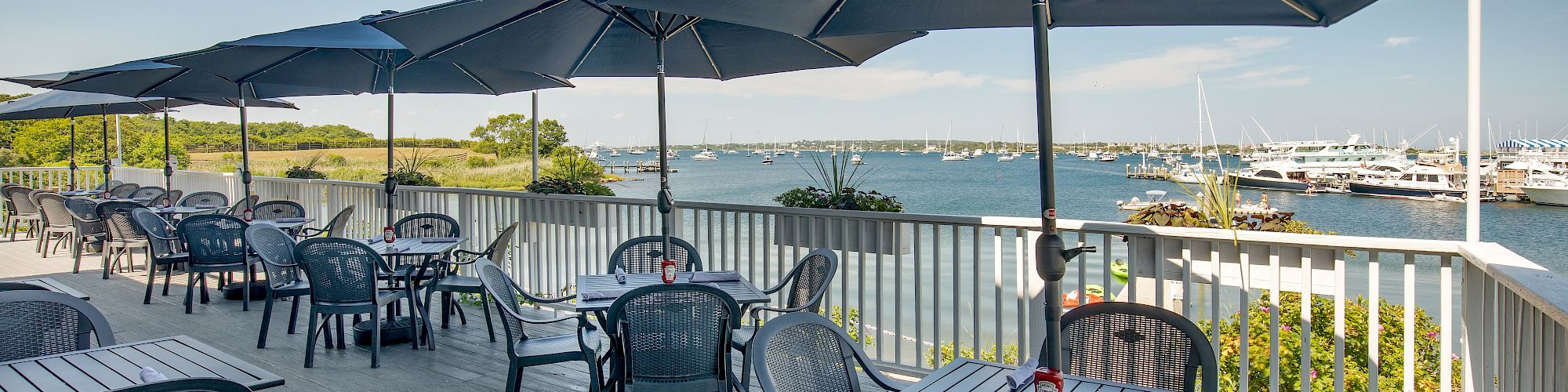 An outdoor dining area with tables, chairs, and blue umbrellas overlooks a scenic waterfront with boats and greenery in the background.