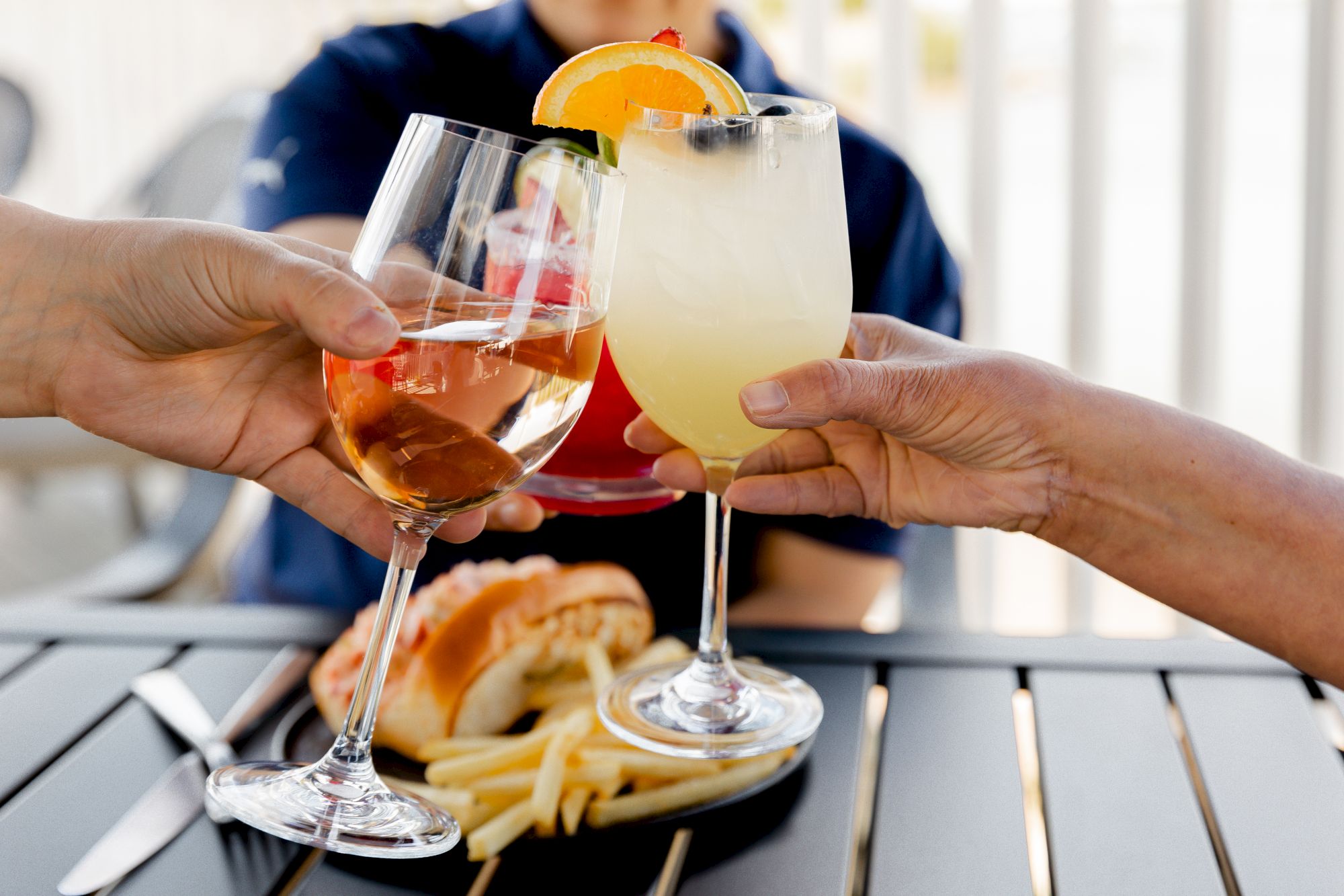 Three glasses with drinks are being clinked together over a table with fries and a sandwich in the background.