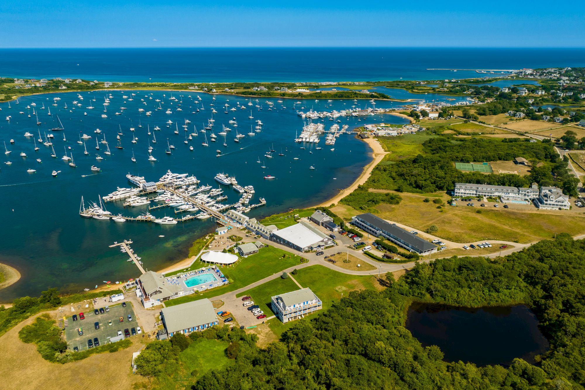 Aerial view of a marina with numerous boats docked, surrounded by buildings, greenery, and a pool, located near the coastline and open sea.