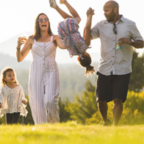 A family of four is enjoying a sunny day outdoors, with the parents playfully swinging one child while the other walks beside them.