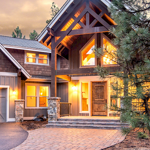 A large, inviting house with warm lighting, surrounded by trees, featuring a spacious driveway and multiple windows, under a twilight sky.