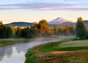 A serene landscape featuring a golf course, a calm river, lush trees, and a snow-capped mountain in the background during sunrise or sunset.