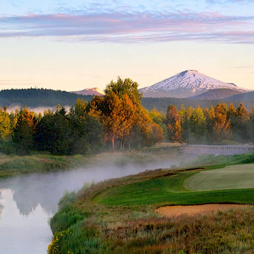A serene landscape featuring a golf course, a calm river, lush trees, and a snow-capped mountain in the background during sunrise or sunset.
