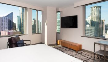 A modern hotel room with large windows showing a cityscape, a wall-mounted TV, a chair, and a wooden cabinet beneath the TV.