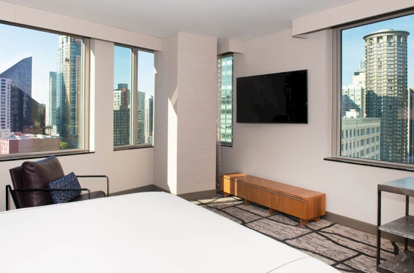 A modern hotel room with large windows showing a cityscape, a wall-mounted TV, a chair, and a wooden cabinet beneath the TV.