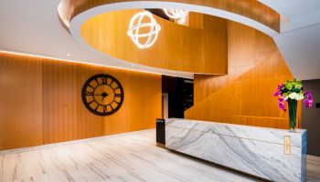 A modern reception area with a wooden spiral staircase, marble desk, wall clock, and flower arrangement on the counter, well-lit and clean.