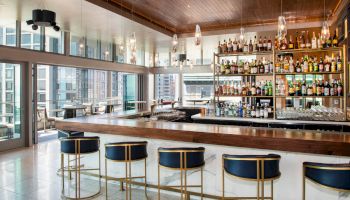 A chic modern bar with stools, a well-stocked bar shelf, and large windows that offer a city view. Glass pendant lights hang from the ceiling.