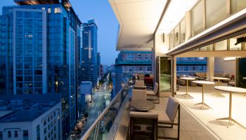 A modern cityscape view from a high balcony at dusk, featuring seating on the terrace, buildings, and a street below, illuminated by streetlights.