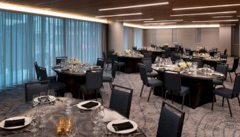 The image shows an elegant dining area set for an event, with round tables, chairs, floral centerpieces, and neatly arranged table settings.