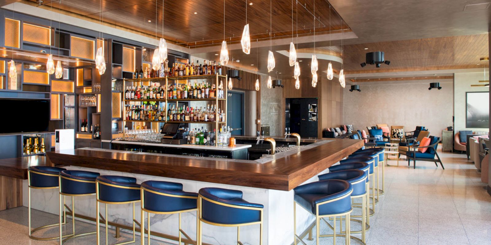 The image shows a modern bar with a wooden counter, blue cushioned stools, hanging lights, and a fully stocked liquor rack behind.
