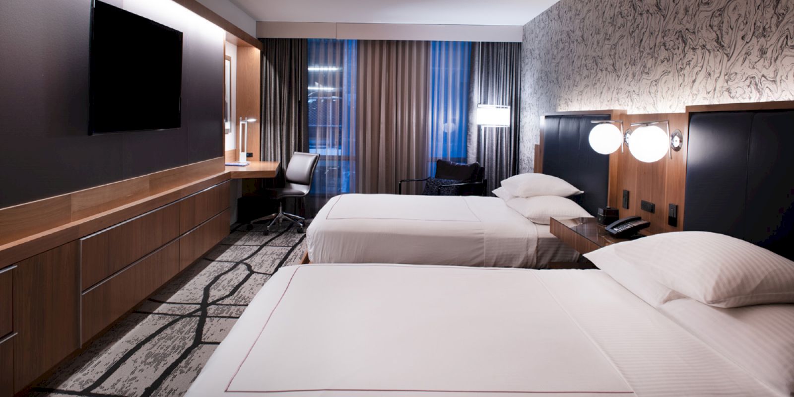 This image shows a modern hotel room with two beds, a TV mounted on the wall, a desk with a chair, and stylish lighting.