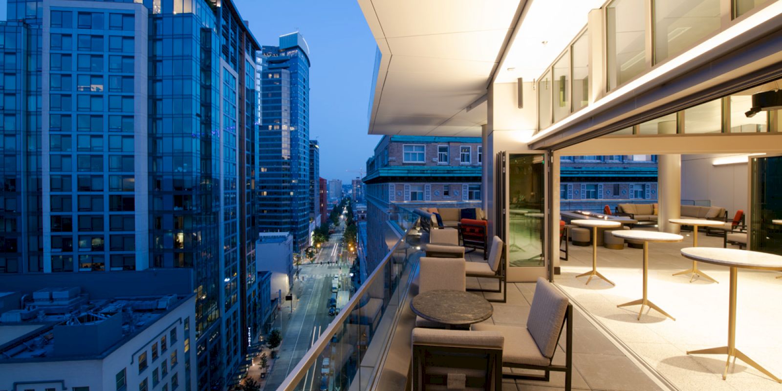 The image shows a modern cityscape with high-rise buildings viewed from a balcony at dusk. The balcony has chairs, tables, and a railing.