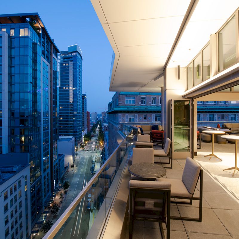 An outdoor balcony with modern furniture overlooks a busy urban street lined with tall buildings during twilight.