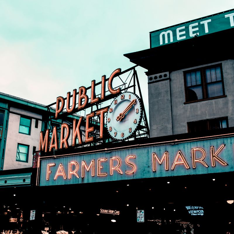 The image shows the entrance to a public market with neon signs reading "Public Market" and "Farmers Market" beneath a clock, surrounded by buildings.