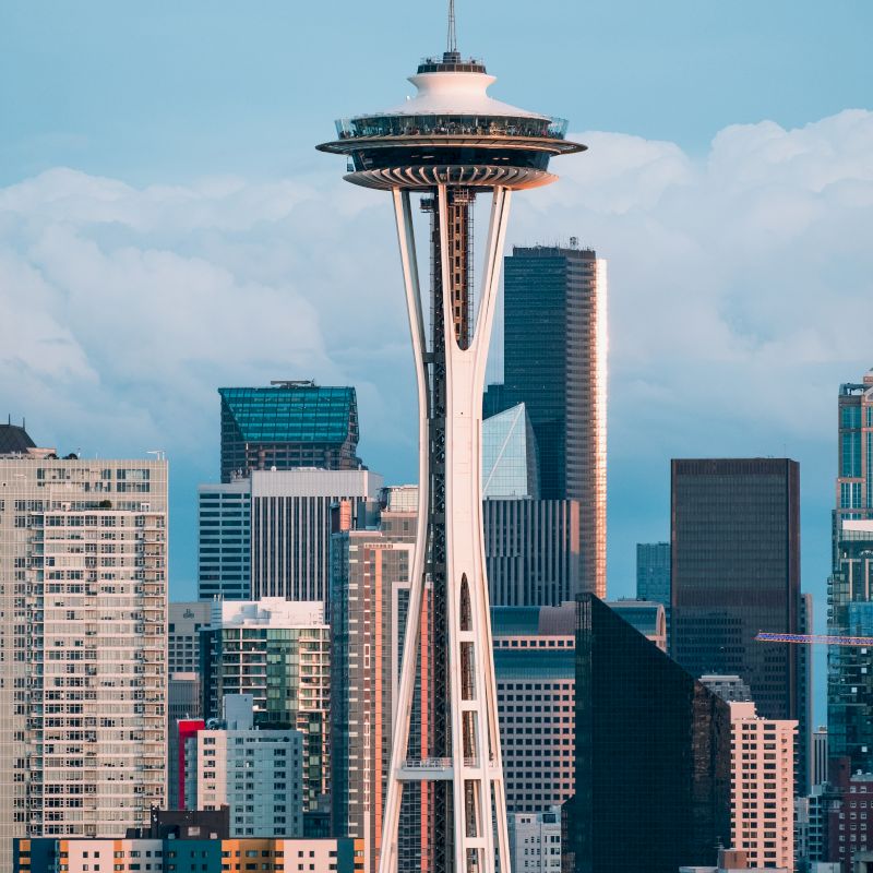 The image shows the Space Needle in Seattle, Washington, with a backdrop of various skyscrapers and buildings under a partly cloudy sky.