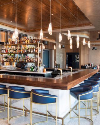 This image shows a modern bar area with a large, well-stocked bar counter, blue and gold bar stools, and stylish lighting fixtures.