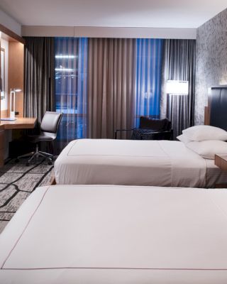A modern hotel room with two beds, a flat-screen TV, a desk, a chair, and a window with curtains; decorated in neutral tones and sleek furnishings.