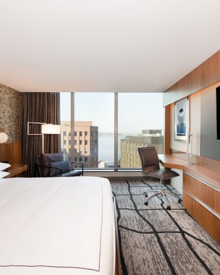 A modern hotel room with a large bed, wall-mounted TV, desk, chair, and window with a city and water view. The decor is contemporary.