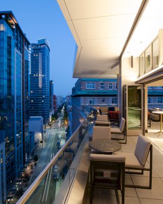 A modern building with an illuminated rooftop terrace featuring seating areas, overlooking a cityscape with high-rise buildings at dusk.