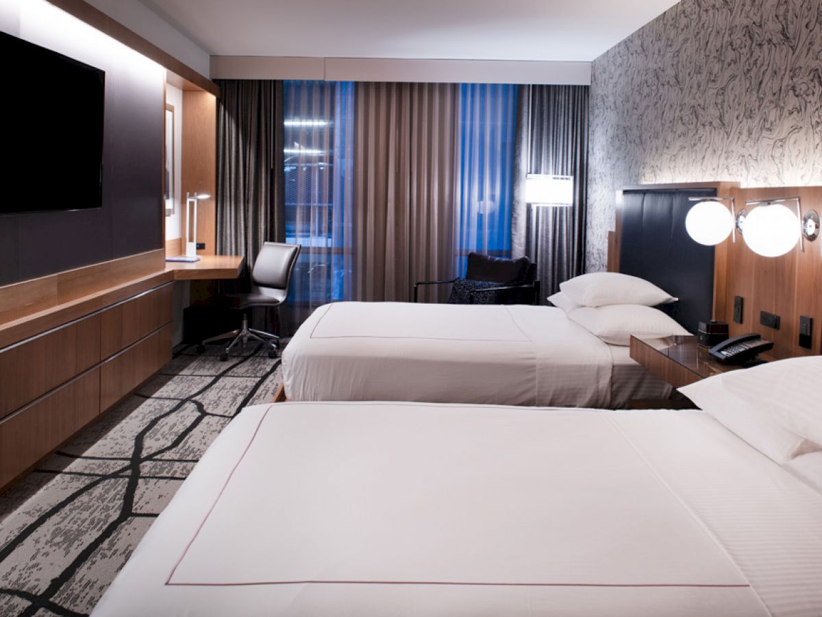 A modern hotel room features two neatly made beds, a flat-screen TV, a work desk, a chair, lamps, and a stylish decor with a large window.