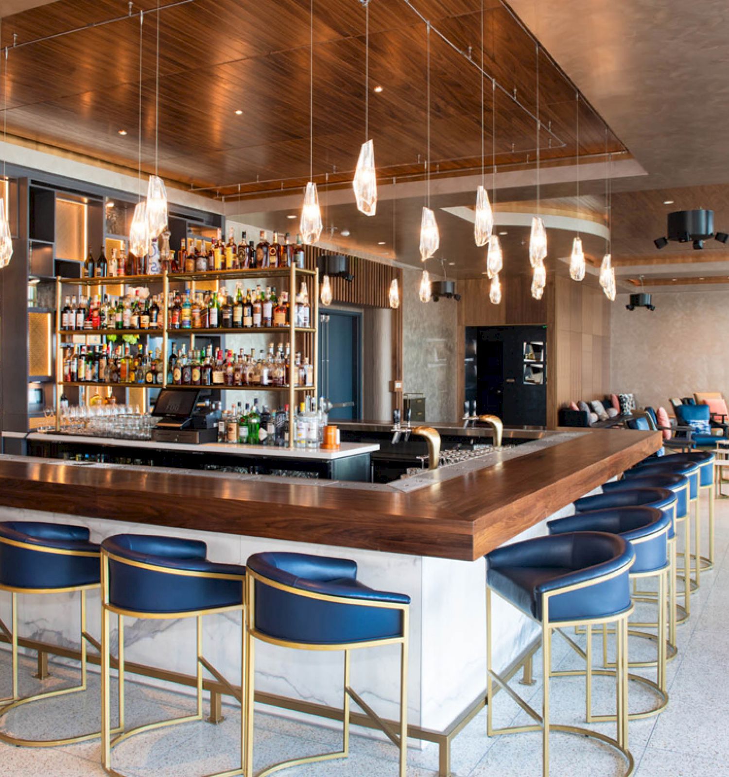 A modern bar area featuring a wooden countertop, blue barstools, hanging lights, and a well-stocked back bar. Lounge seating is visible in the background.