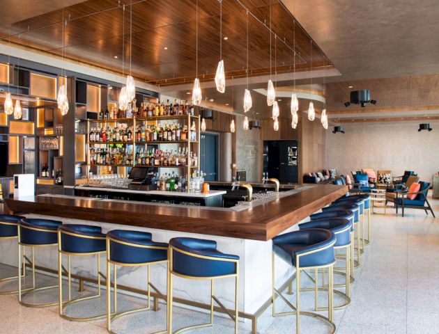 A modern bar with a wooden counter, blue bar stools, hanging lights, and a wide selection of bottles on display. The seating area is in the background.