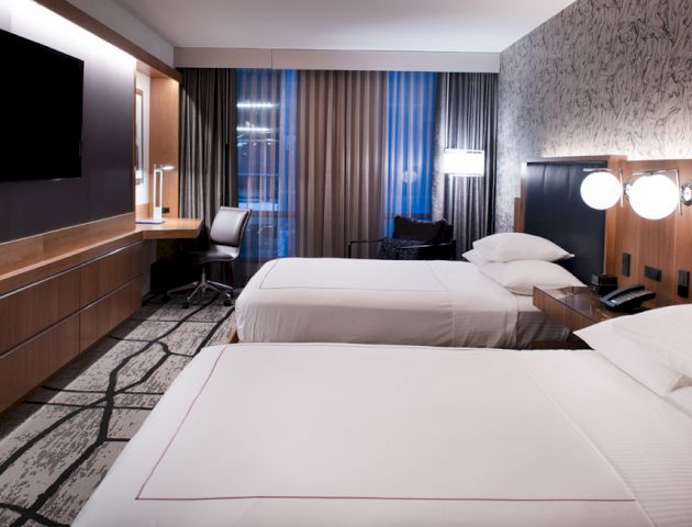 This image shows a modern hotel room with two beds, a flat-screen TV, a desk, and a chair by the window, featuring contemporary decor and lighting.