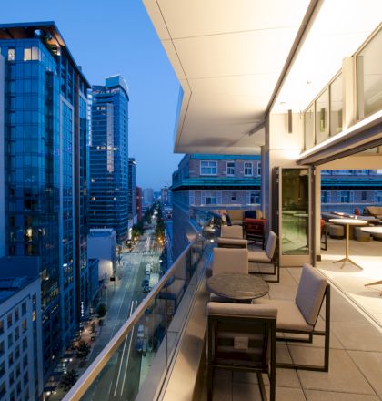A modern balcony with city views at dusk, featuring tables and seating. Adjacent buildings and street below are also visible.
