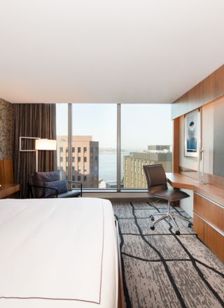 This image shows a modern hotel room with a large bed, floor-to-ceiling windows, a desk with a chair, a flat-screen TV, and a view of buildings.