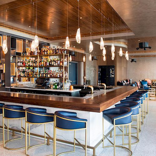 A modern bar features a sleek counter, blue bar stools, hanging lights, and a well-stocked shelf. Cozy seating areas are in the background.