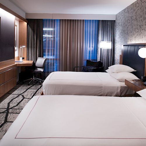 This image shows a modern hotel room with two double beds, bedside tables, a desk with a chair, a TV, and patterned wallpaper.