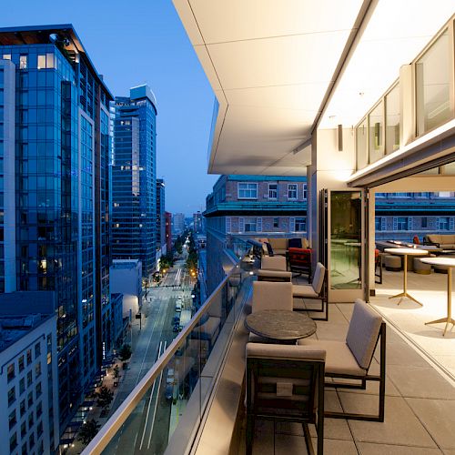 An outdoor terrace with tables and chairs overlooks a cityscape with modern high-rise buildings at dusk, featuring a vibrant urban atmosphere.