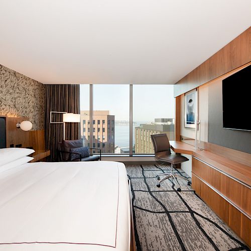A modern hotel room features a large bed, a desk with a chair, a wall-mounted TV, and a view of buildings outside the large window.