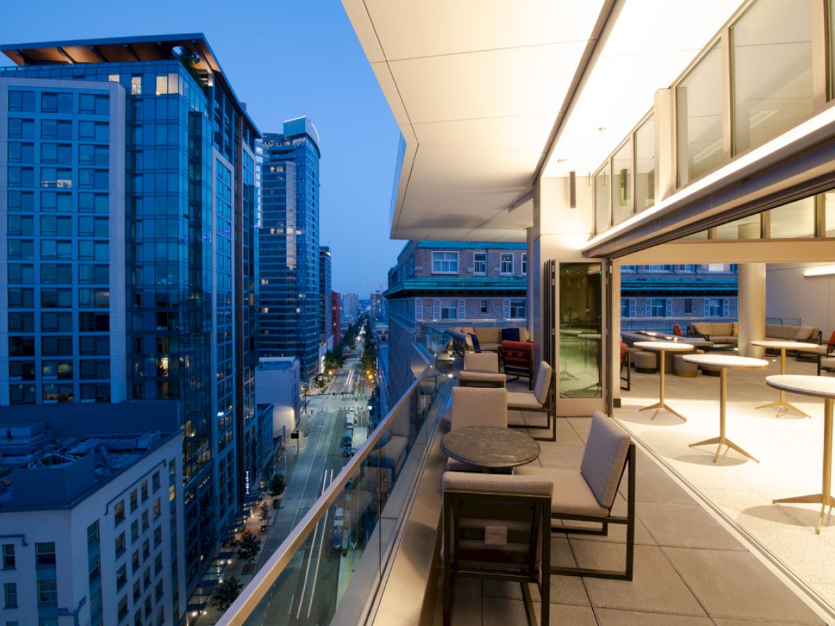 A modern cityscape at dusk with high-rise buildings and a spacious balcony featuring outdoor seating and tables.