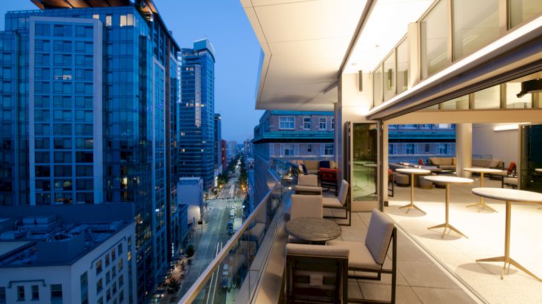 A modern cityscape at dusk with high-rise buildings and a spacious balcony featuring outdoor seating and tables.