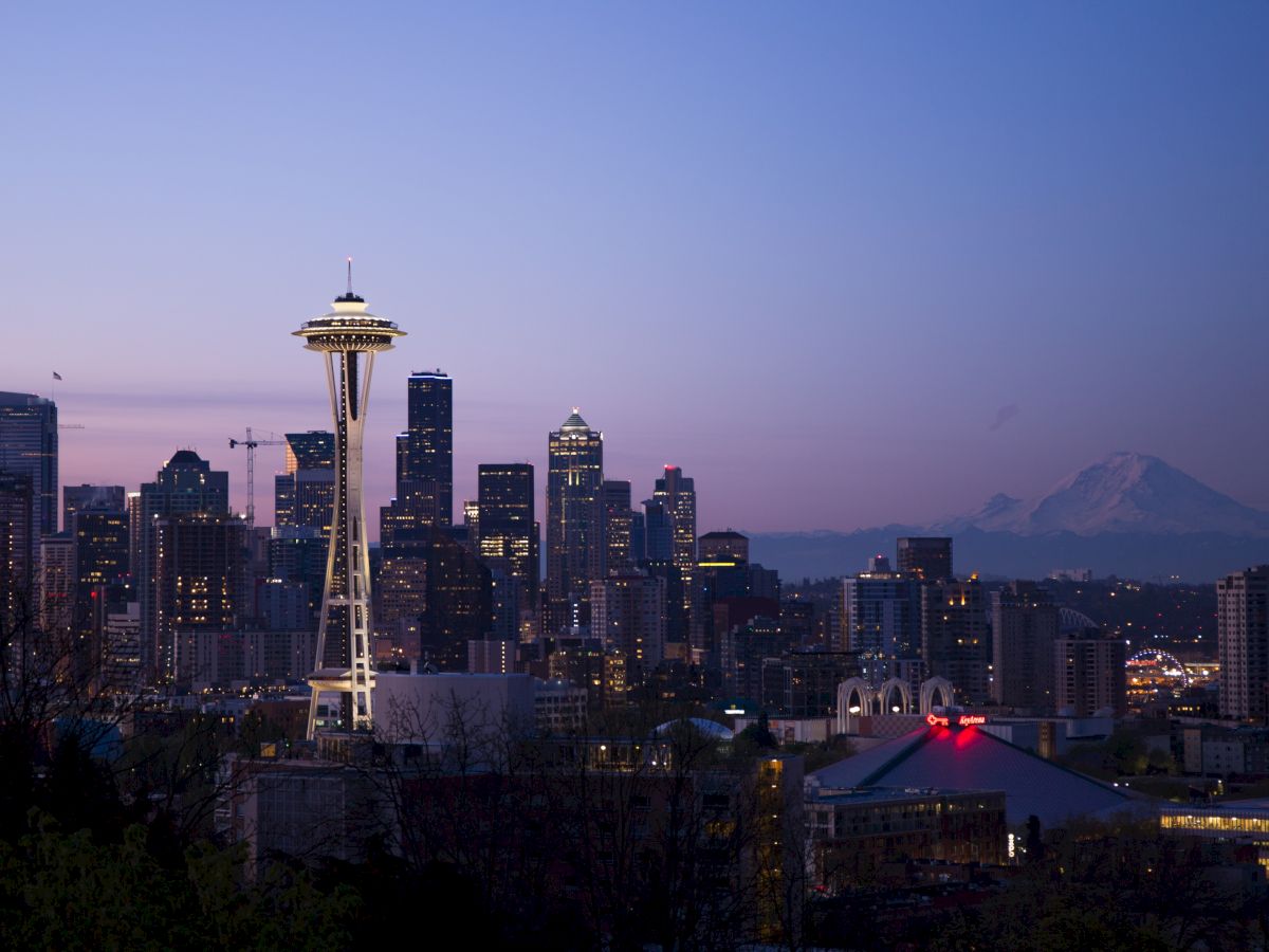 This image shows a city skyline at dusk, featuring a prominent tower, numerous skyscrapers, and a distant mountain range under a twilight sky.