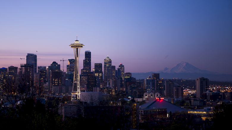This image shows a city skyline at dusk, featuring a prominent tower, numerous skyscrapers, and a distant mountain range under a twilight sky.