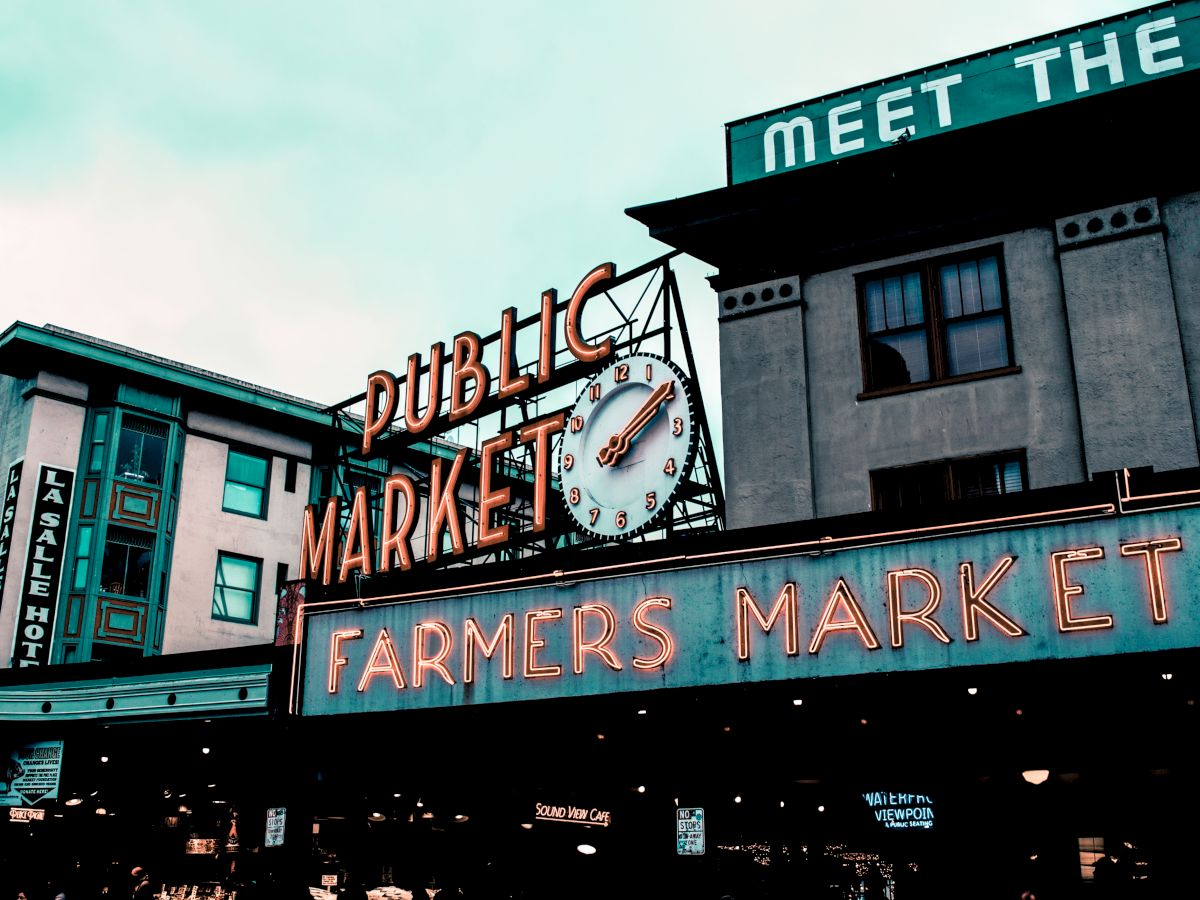 The image shows the iconic Public Market Center sign with a clock and the Farmers Market sign in neon lights, likely Pike Place Market in Seattle, WA.