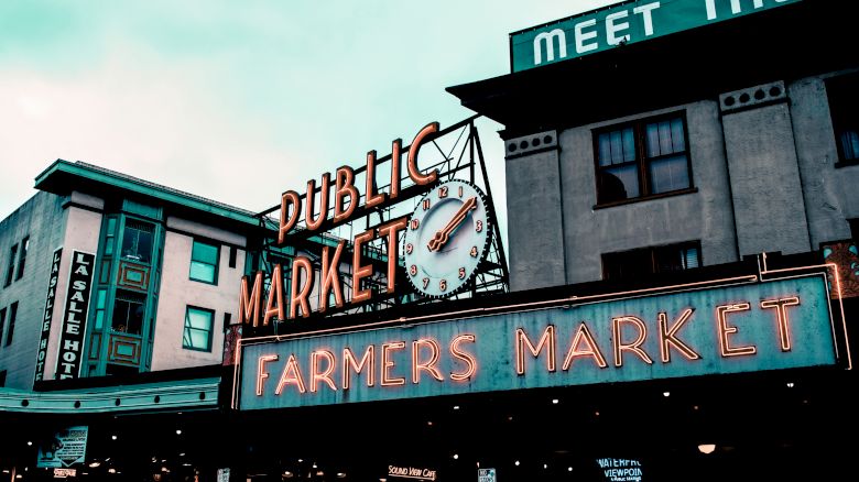 The image shows the iconic Public Market Center sign with a clock and the Farmers Market sign in neon lights, likely Pike Place Market in Seattle, WA.