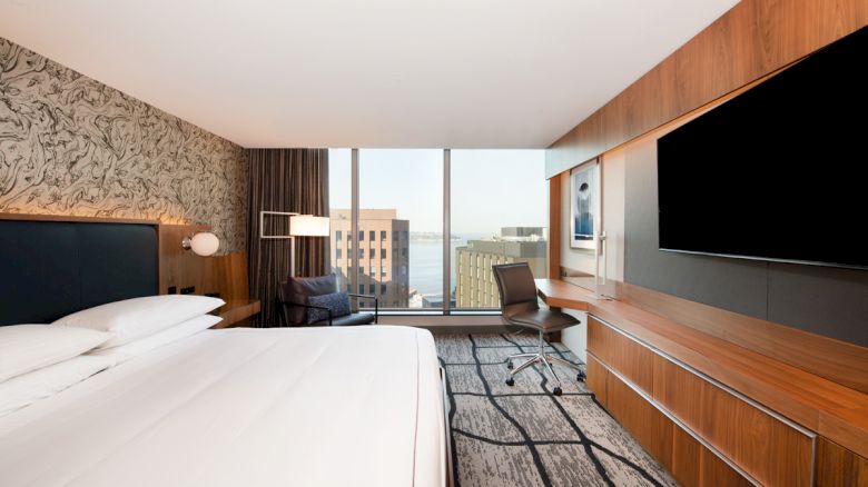 A modern hotel room with a large bed, a desk with a chair, a mounted TV, and a view of buildings and the river through a large window.