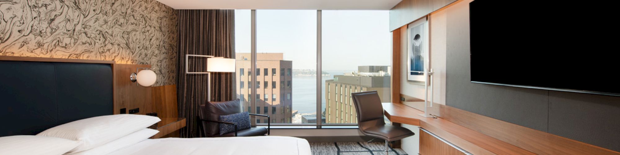 A modern hotel room with a large bed, wall-mounted TV, desk, and chair. It has a city view from the large windows.