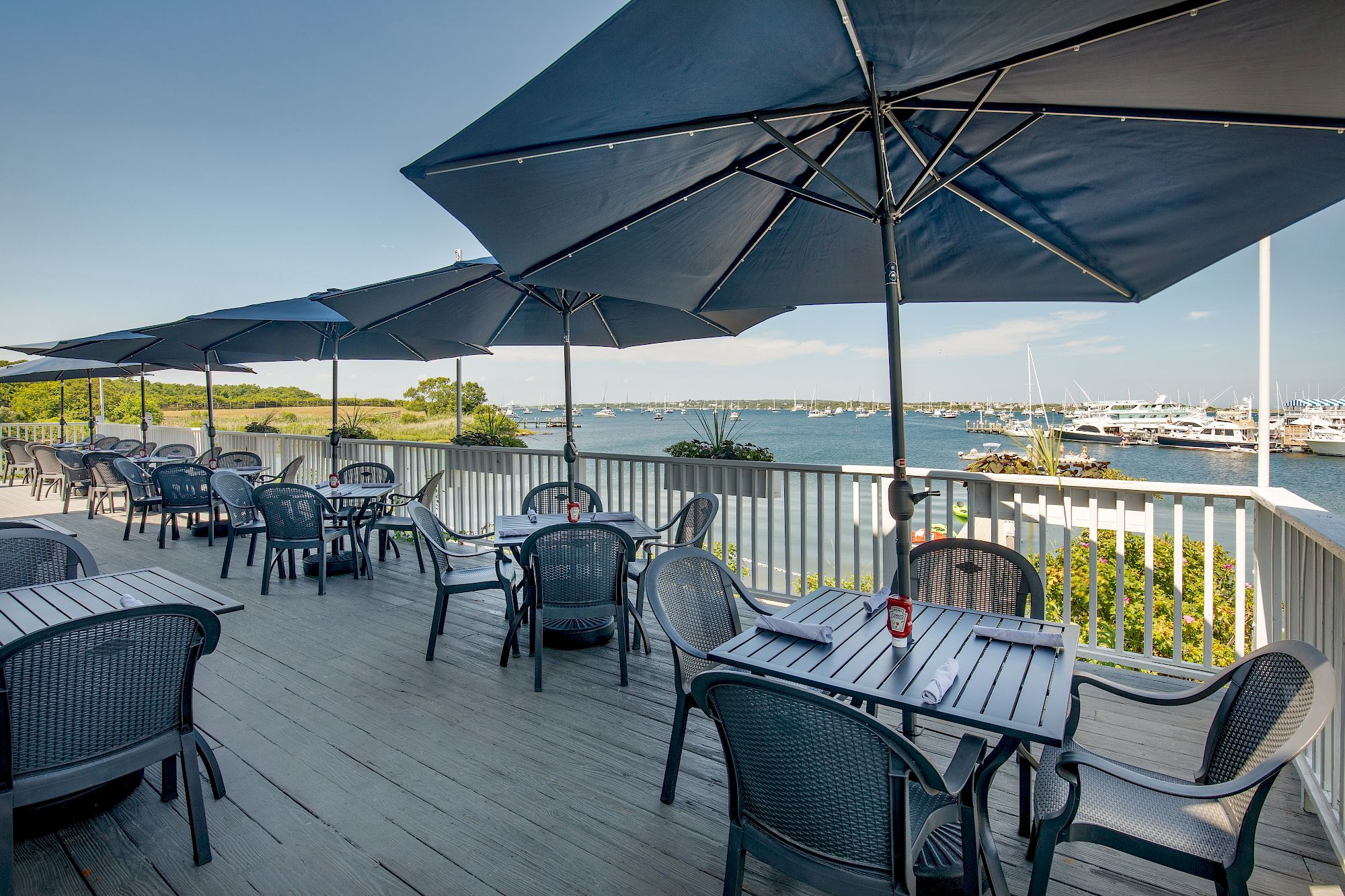 An outdoor dining area with tables and chairs is shaded by large umbrellas overlooking a scenic water view with boats and greenery in the background.