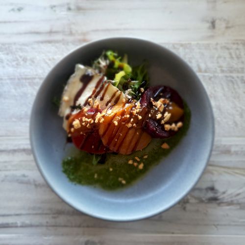 A grey bowl with a colorful salad including sliced beets, greens, sauce, and a sprinkle of nuts on a white wooden table.