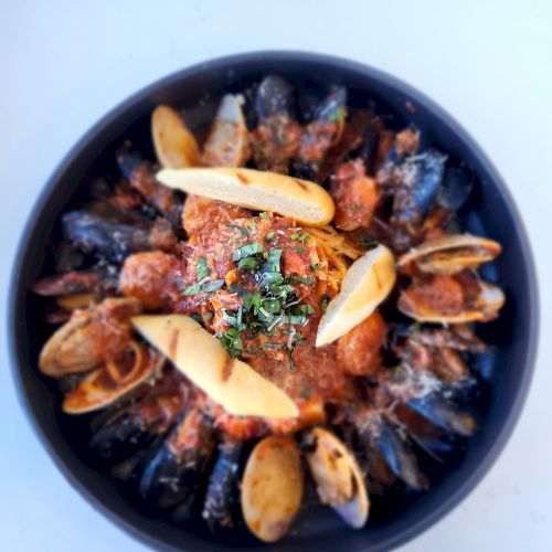 The image shows a bowl of seafood stew garnished with herbs and pieces of toasted bread on top, served in a black bowl.