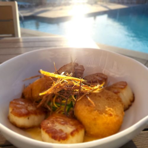 A bowl of scallops and croquettes garnished with thinly sliced vegetables, placed on an outdoor table near a pool with the sun setting.