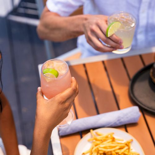 Two people enjoying drinks with lime and a meal of fries and a sandwich at an outdoor restaurant.