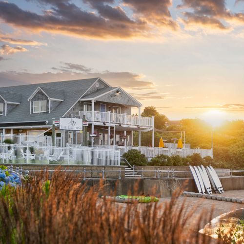 A beautiful house by the waterside at sunset, with a wooden dock, lush greenery, and a dramatic sky with clouds.