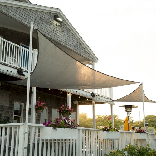 A white porch with railings and sunshades, featuring hanging plants and an outdoor heater, is attached to a house.